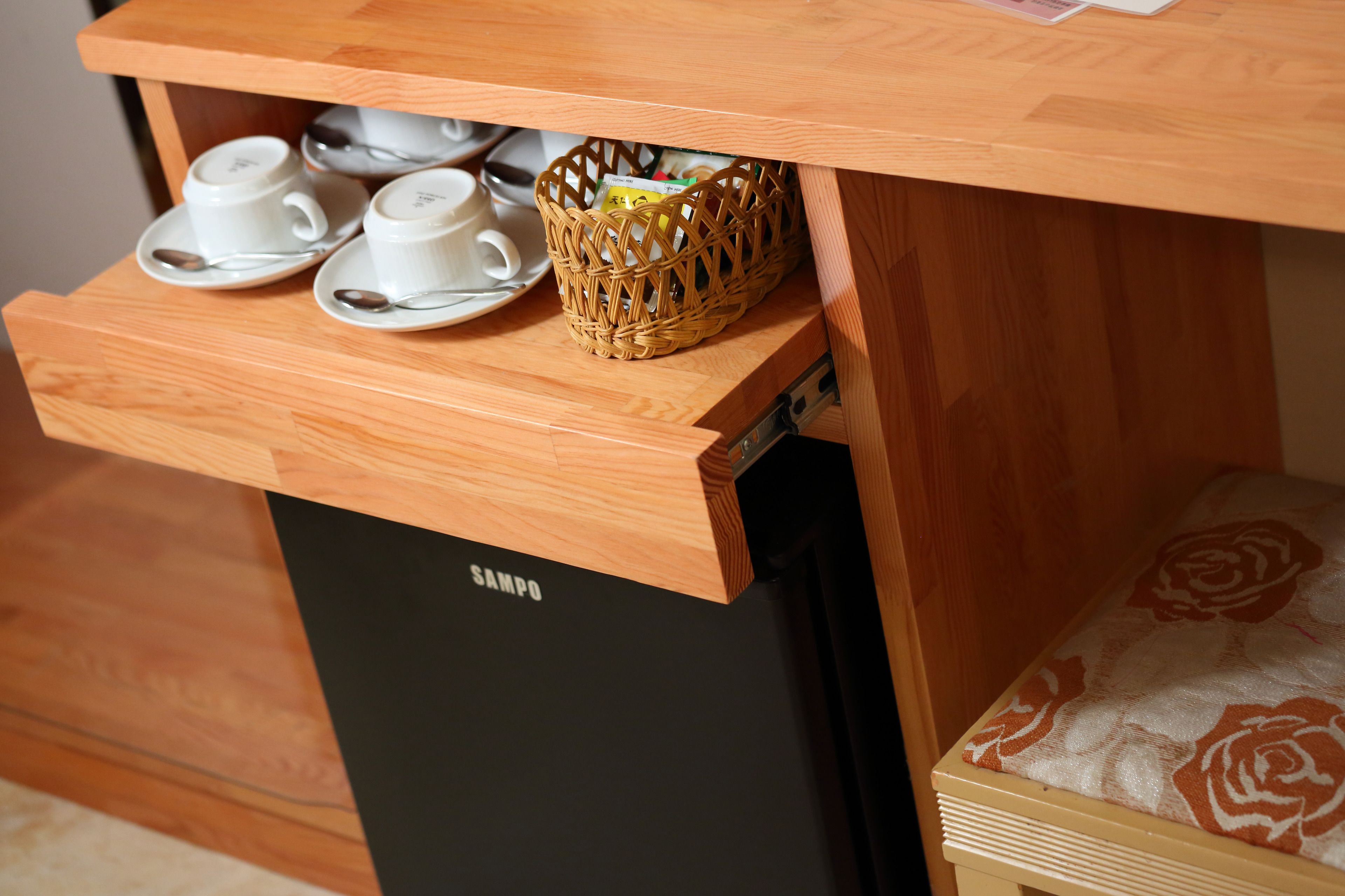 A wooden cabinet with a drawer containing white cups and saucers, a wicker basket, and a black appliance labeled "SAMPO".