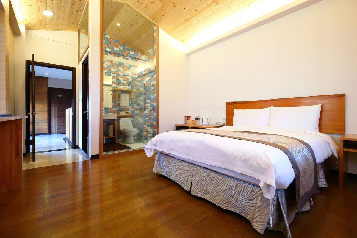 A modern hotel room with a wooden floor, a large bed with a zebra-patterned skirt, a wooden headboard, and a glass shower enclosure with a mosaic tile wall.