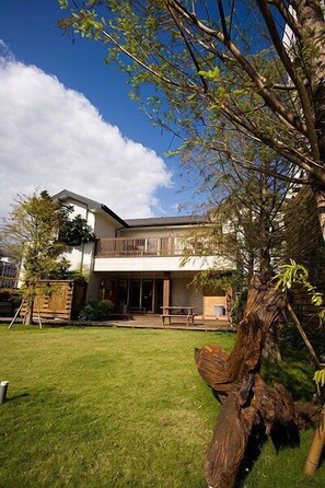 Una casa moderna de dos pisos con un amplio jardín delantero, una terraza de madera y un gran árbol en primer plano.
