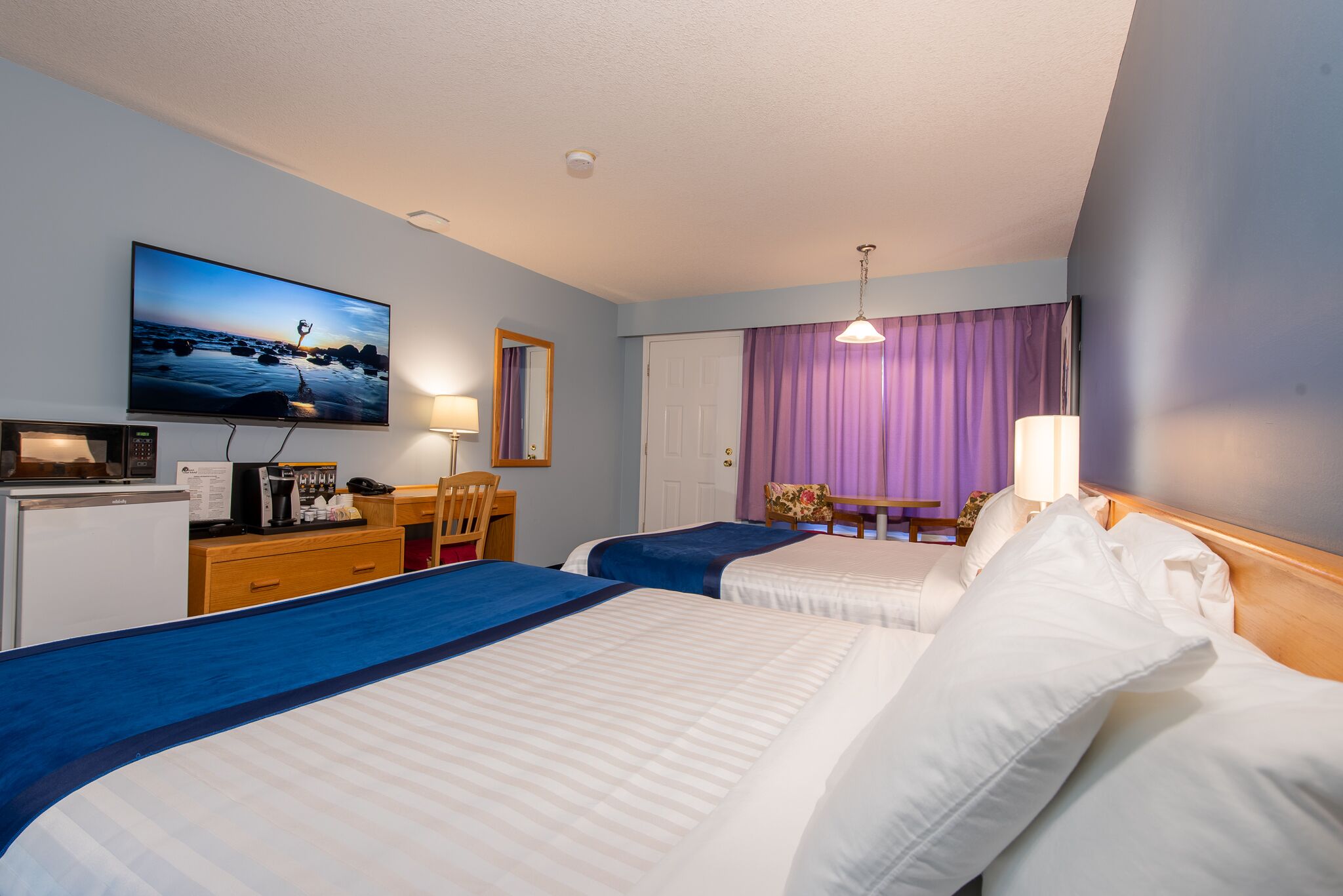 Habitación de hotel con dos camas, un escritorio con computadora, televisor montado en la pared y una ventana con cortinas moradas.