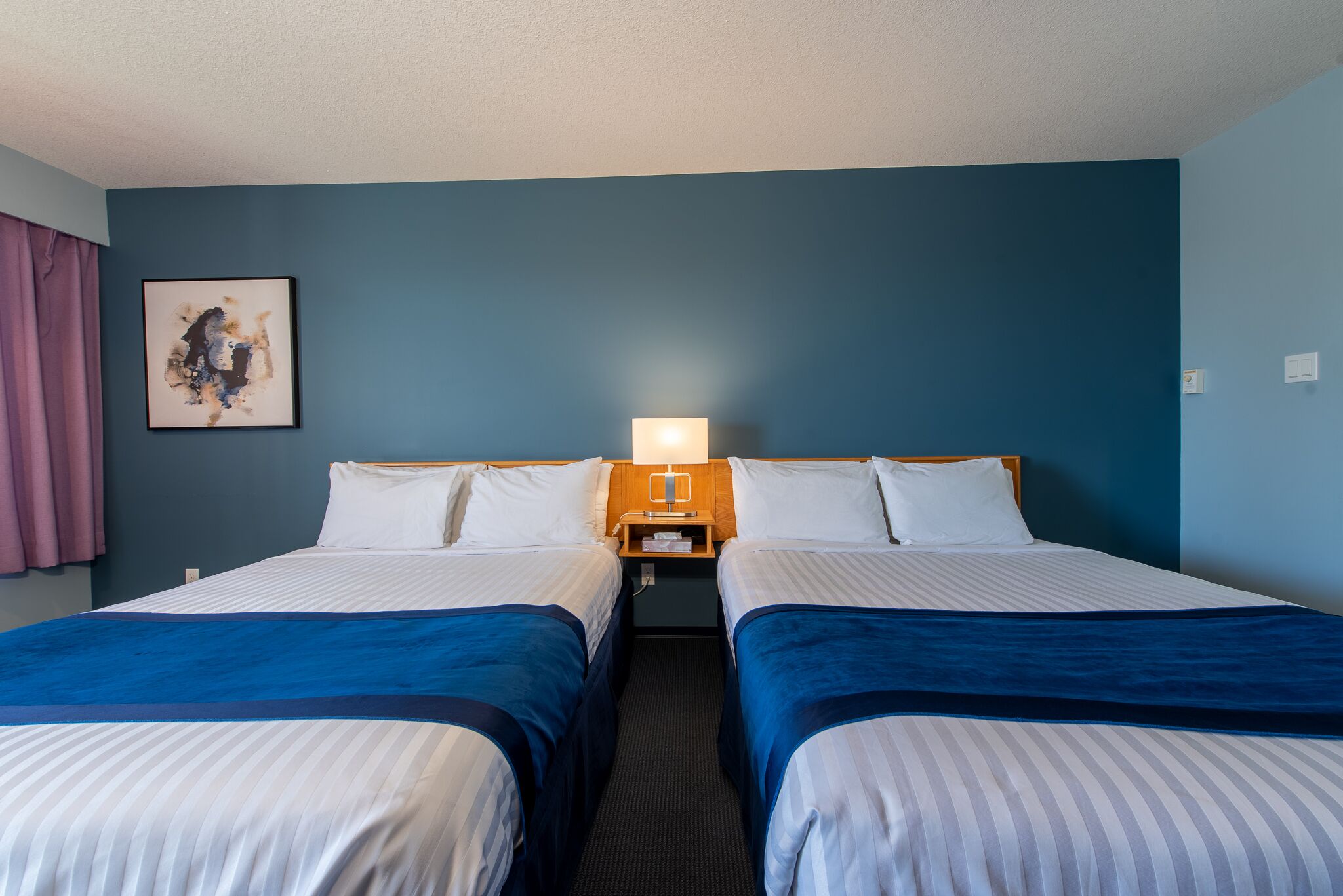 A hotel room with two beds, a nightstand with a lamp, a painting on the wall, and a window with curtains.