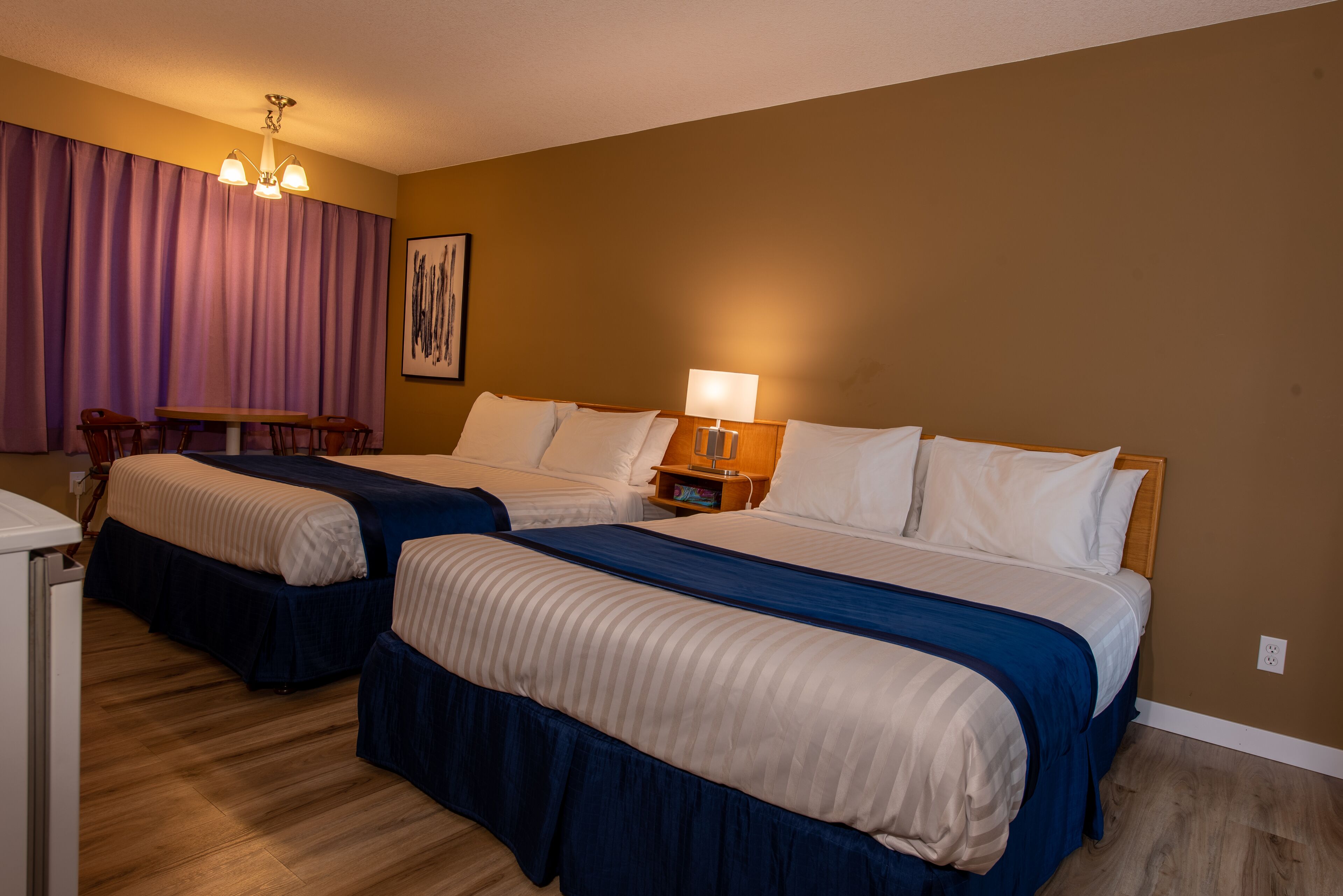 A hotel room with two beds, a desk, a chair, a lamp, and a painting on the wall.