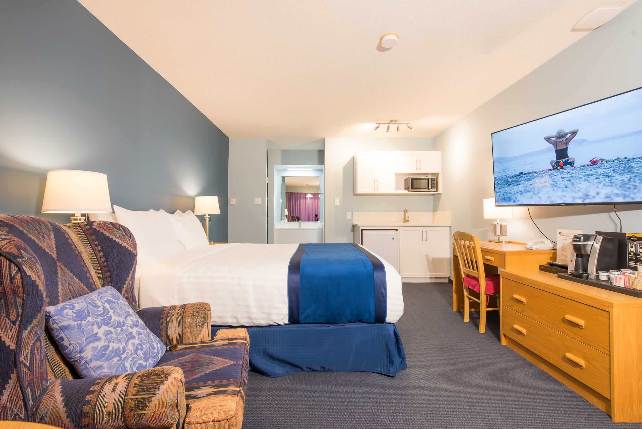 A hotel room with a bed, a sofa, a desk, a television, and a kitchenette.