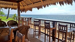 A seaside restaurant with thatched roof, wooden tables, and wicker chairs, overlooking the ocean.