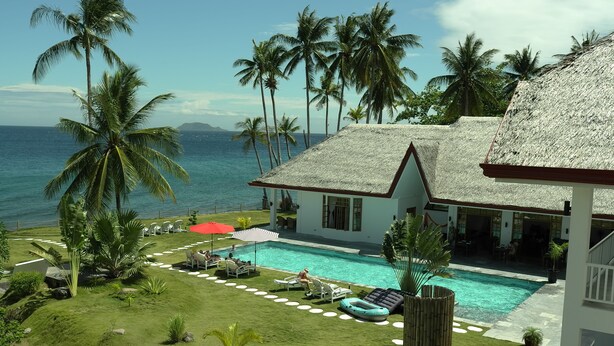 A pool area with lounge chairs and umbrellas, surrounded by palm trees and a view of the ocean.