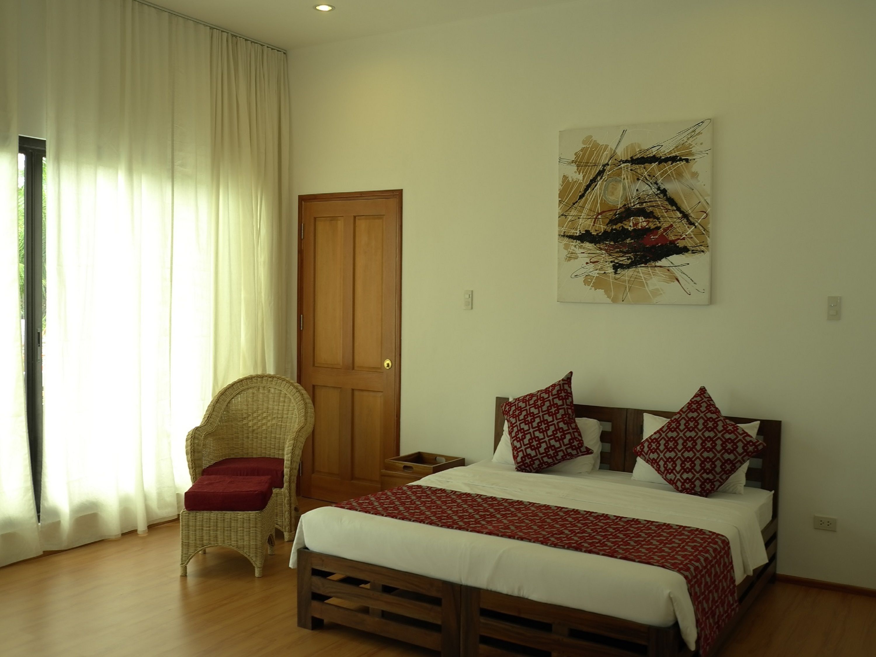 Quarto com cama de madeira, cadeira de vime, porta de madeira e um quadro na parede.