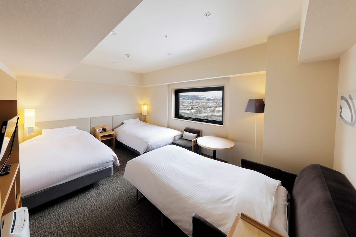 A hotel room with three beds, a small round table, a sofa, and a window with a city view.