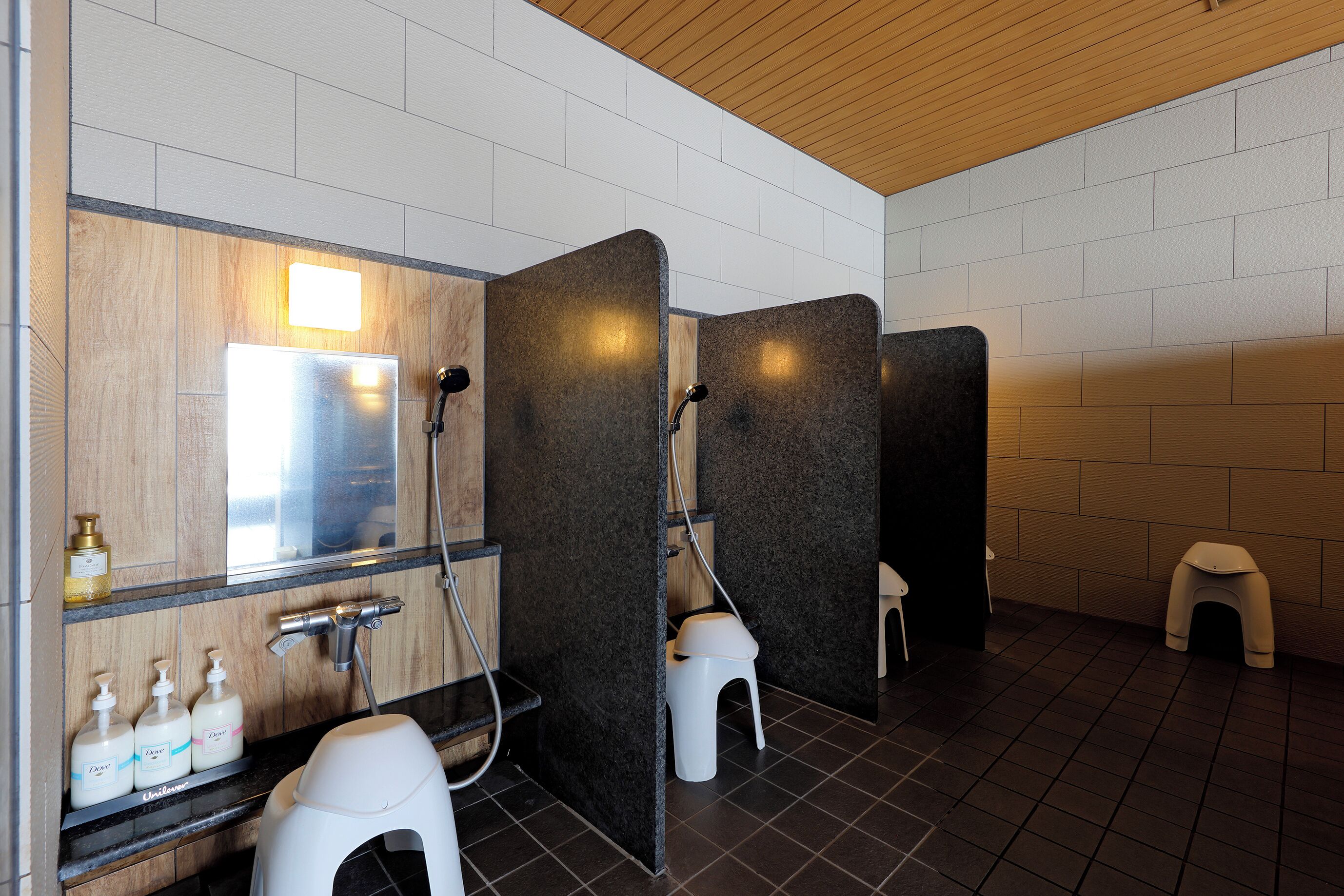 A modern bathroom with three individual shower stalls, a vanity with soap dispensers, and a wooden ceiling.
