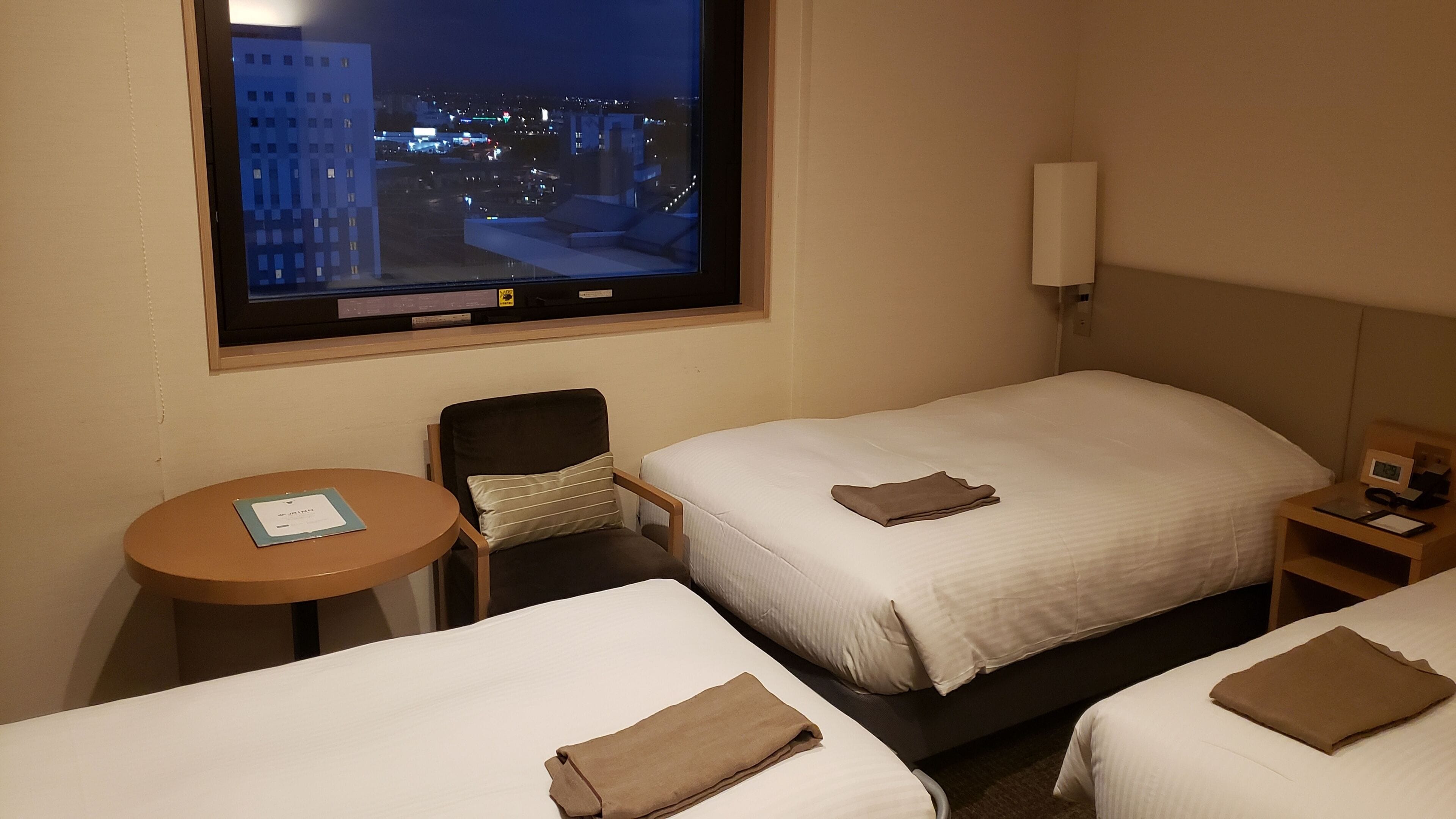 A hotel room with two beds, a small table, a chair, a window with a city view, and a nightstand with a lamp.