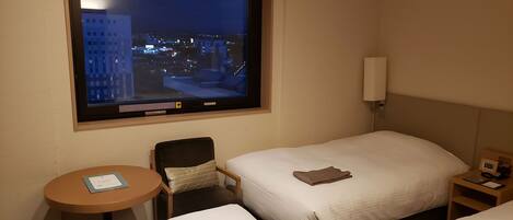 A hotel room with two beds, a small table, a chair, a window with a city view, and a nightstand with a lamp.