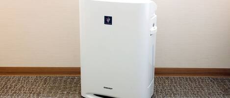 A white portable air conditioner in a corner with a wooden baseboard and a textured carpet.