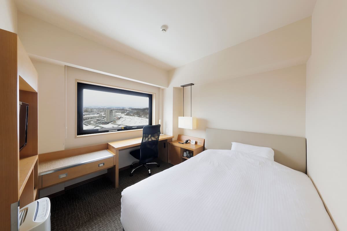 A hotel room with a bed, a desk, a chair, a window with a view, and a bedside table with a lamp.