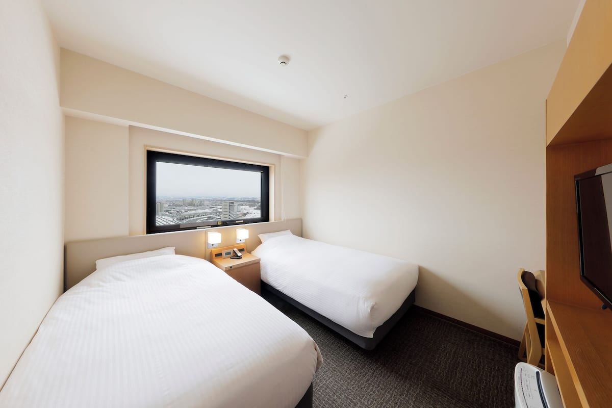 A hotel room with two beds, a window with a city view, a small bedside table with a lamp, and a television on the right.