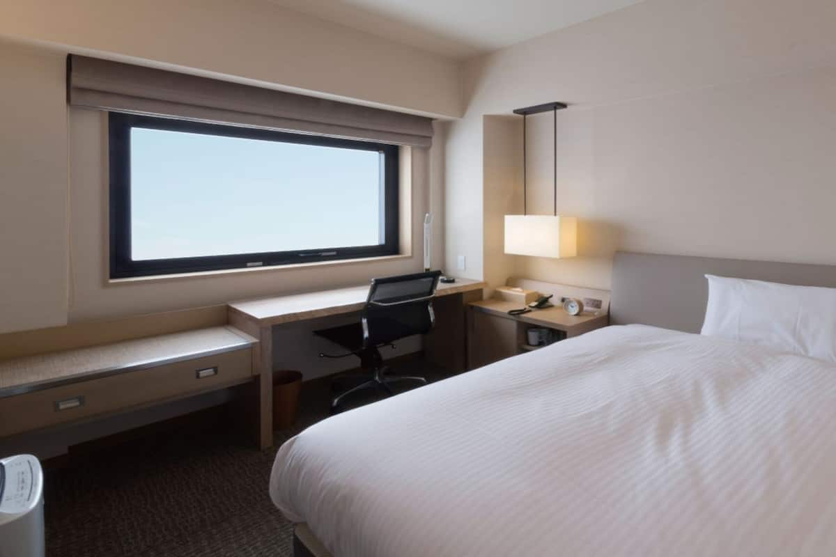 A hotel room with a bed, desk, chair, lamp, and window.