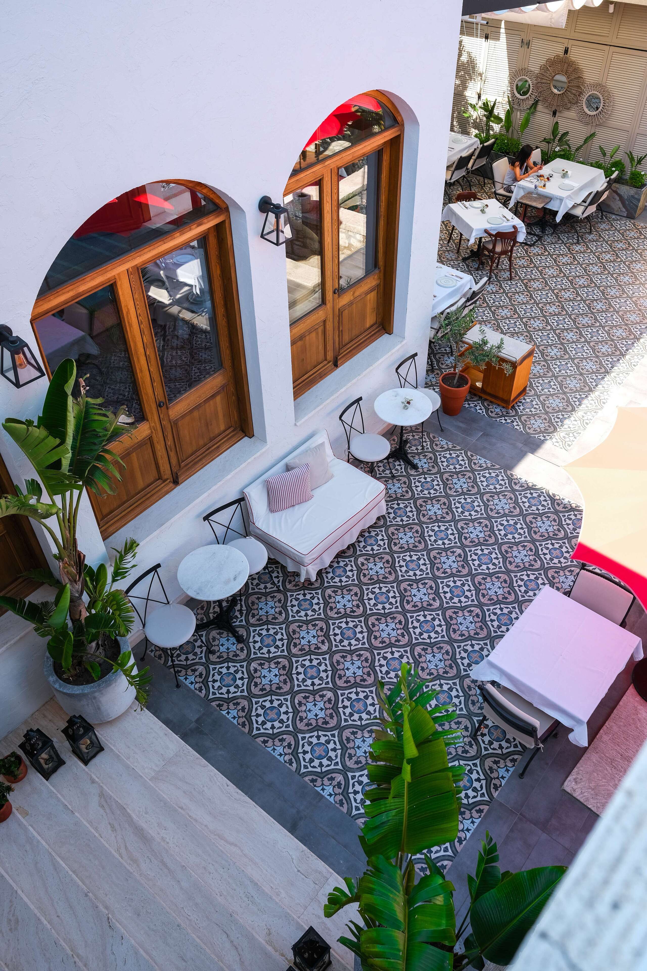 An outdoor patio with a tiled floor, white walls, wooden-framed windows, and various seating arrangements including tables and chairs.