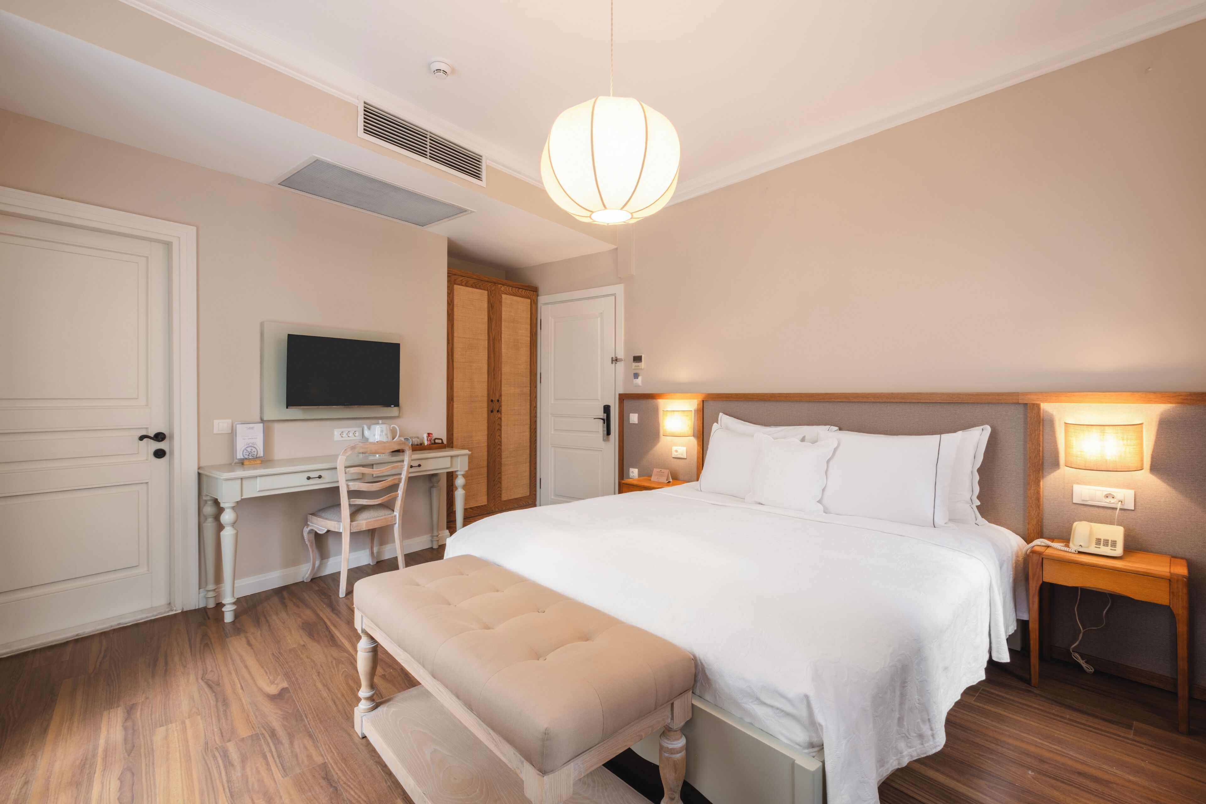 A hotel room with a bed, a desk, a chair, a television, and a bedside table with a lamp.