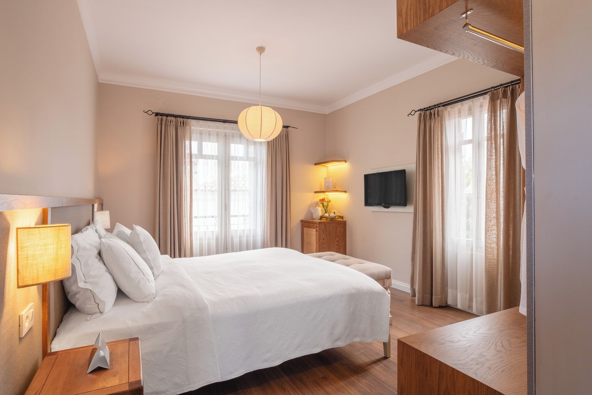 Habitación de hotel con cama, lámpara de noche, mesita de noche, banco de madera, cortinas y televisor montado en la pared.