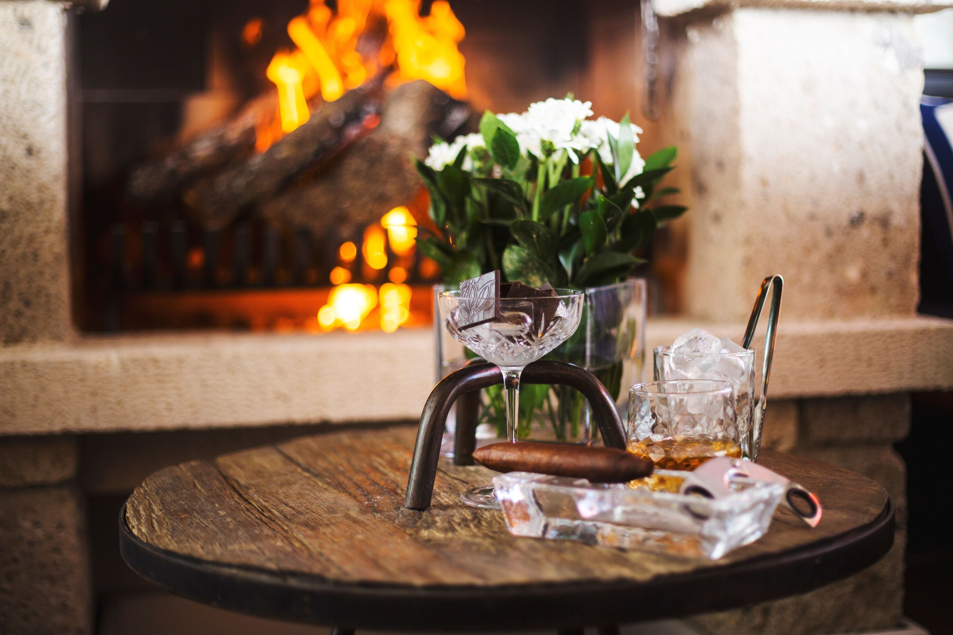 A round wooden table with a glass, a cigar, and ice cubes. In the background, there is a fireplace with a log and white flowers.