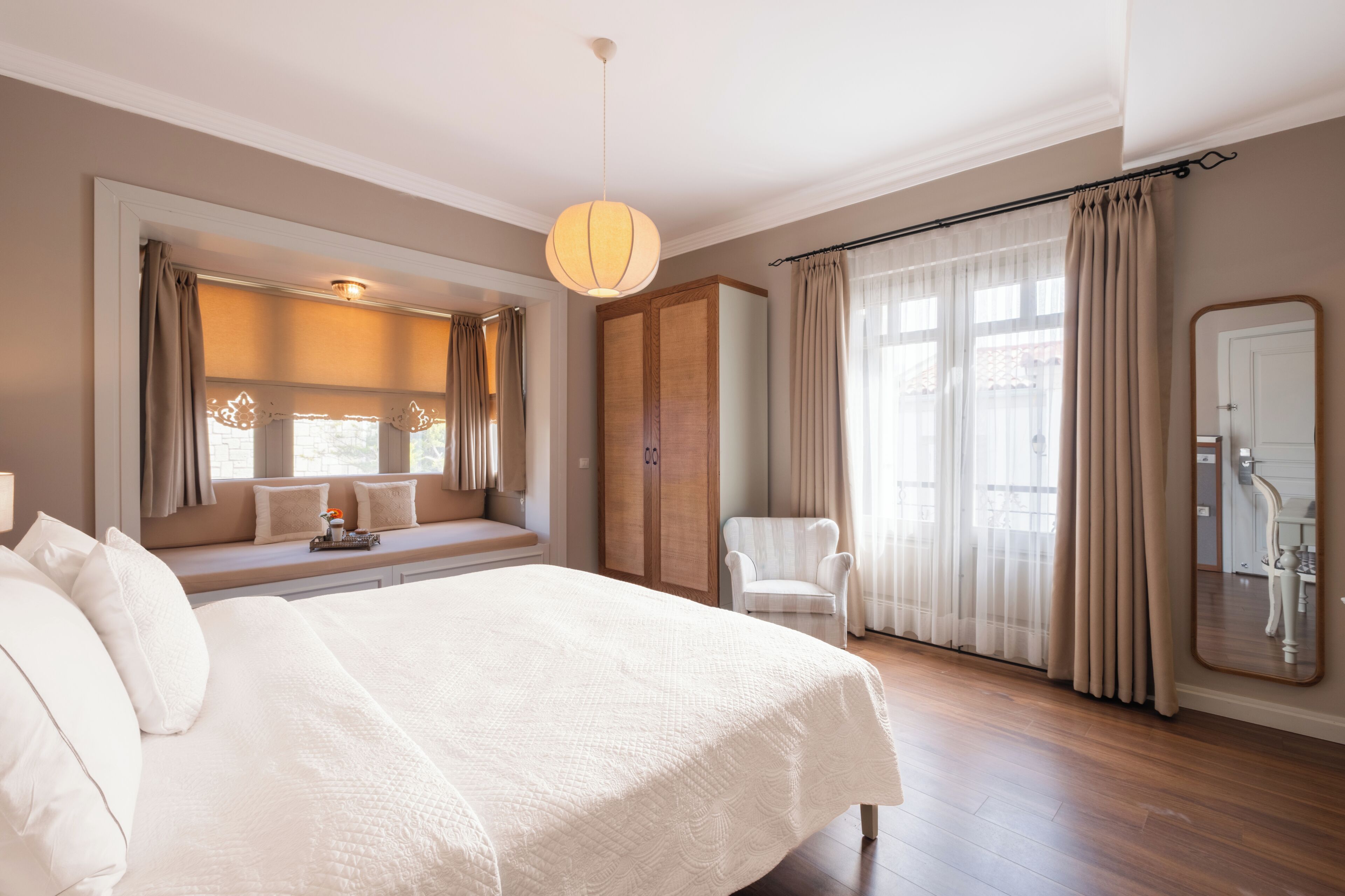 A bedroom with a large bed, a window seat, a chair, a wardrobe, and a mirror.