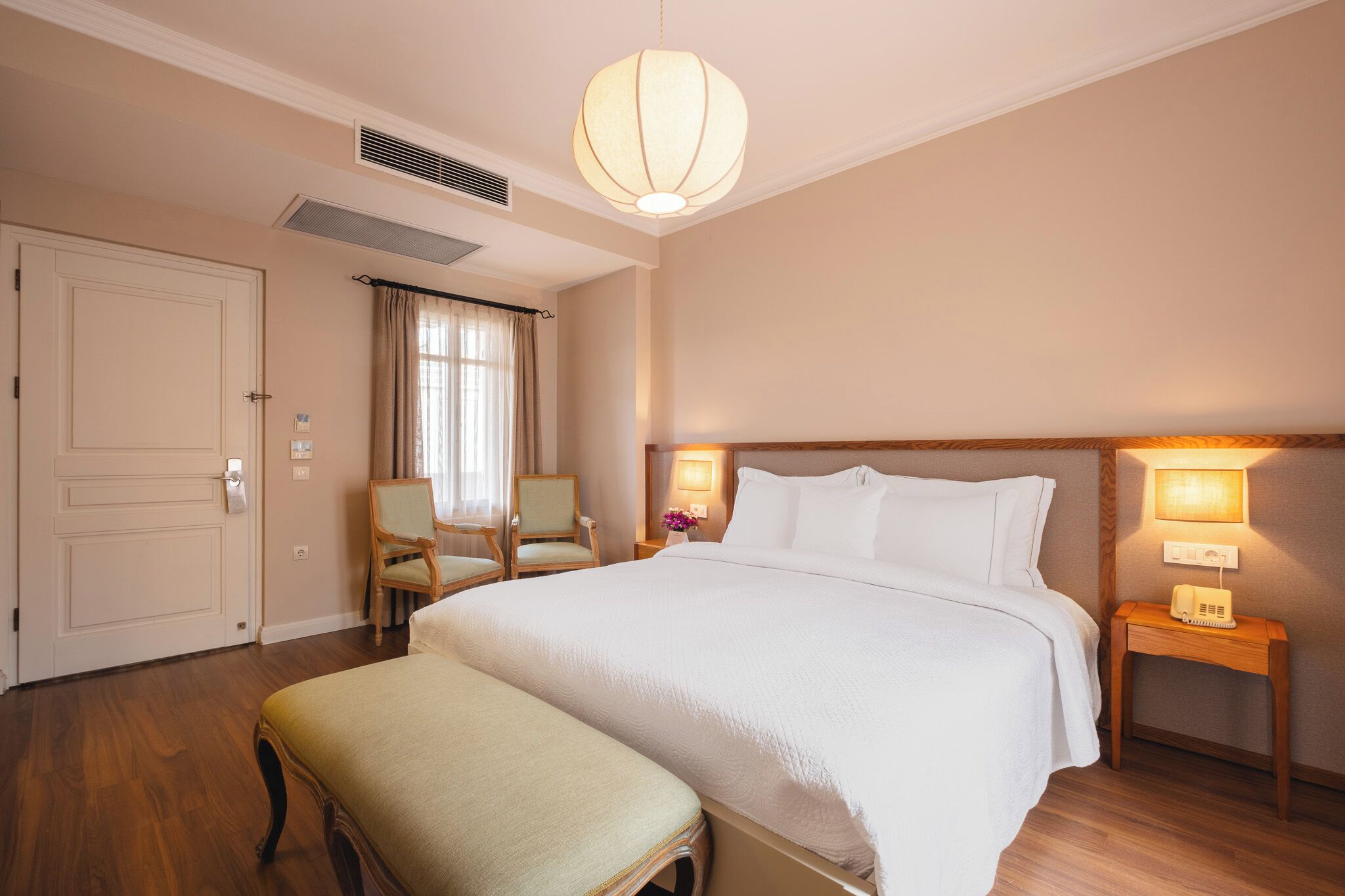 A hotel room with a large bed, a bedside table with a lamp, a chair, and a window with curtains.