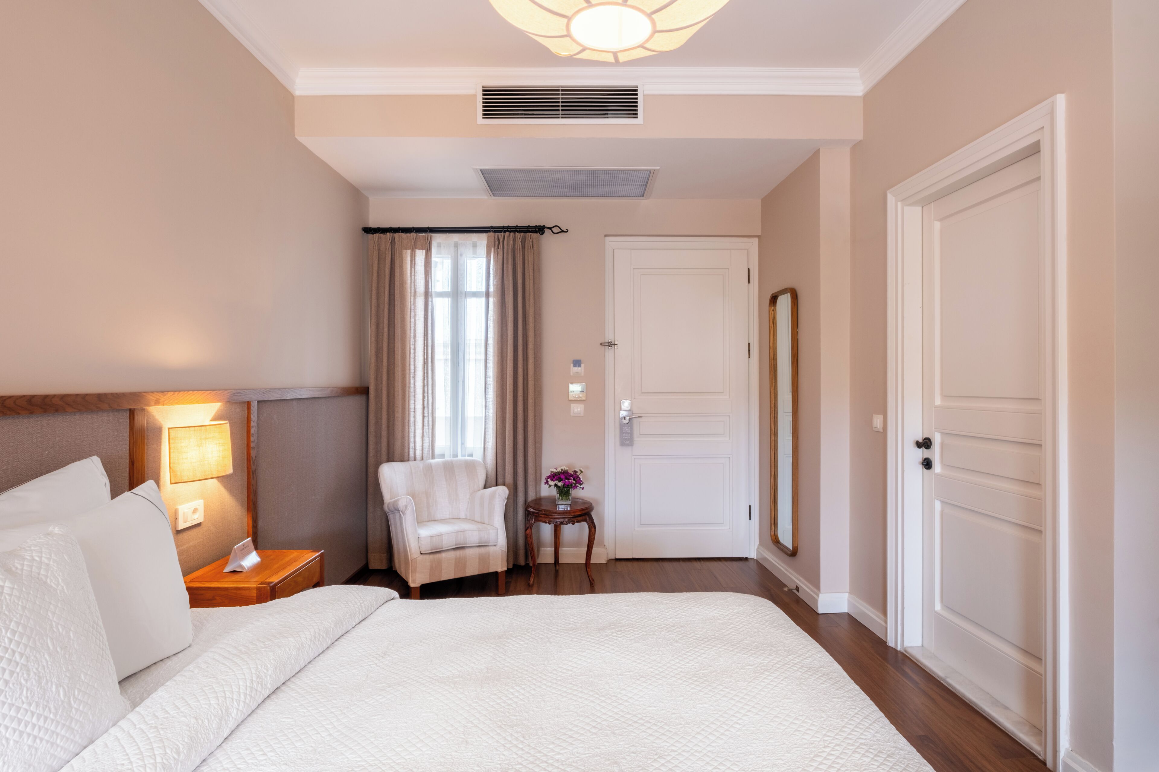 A hotel room with a bed, a chair, a nightstand, a lamp, a mirror, and a door.