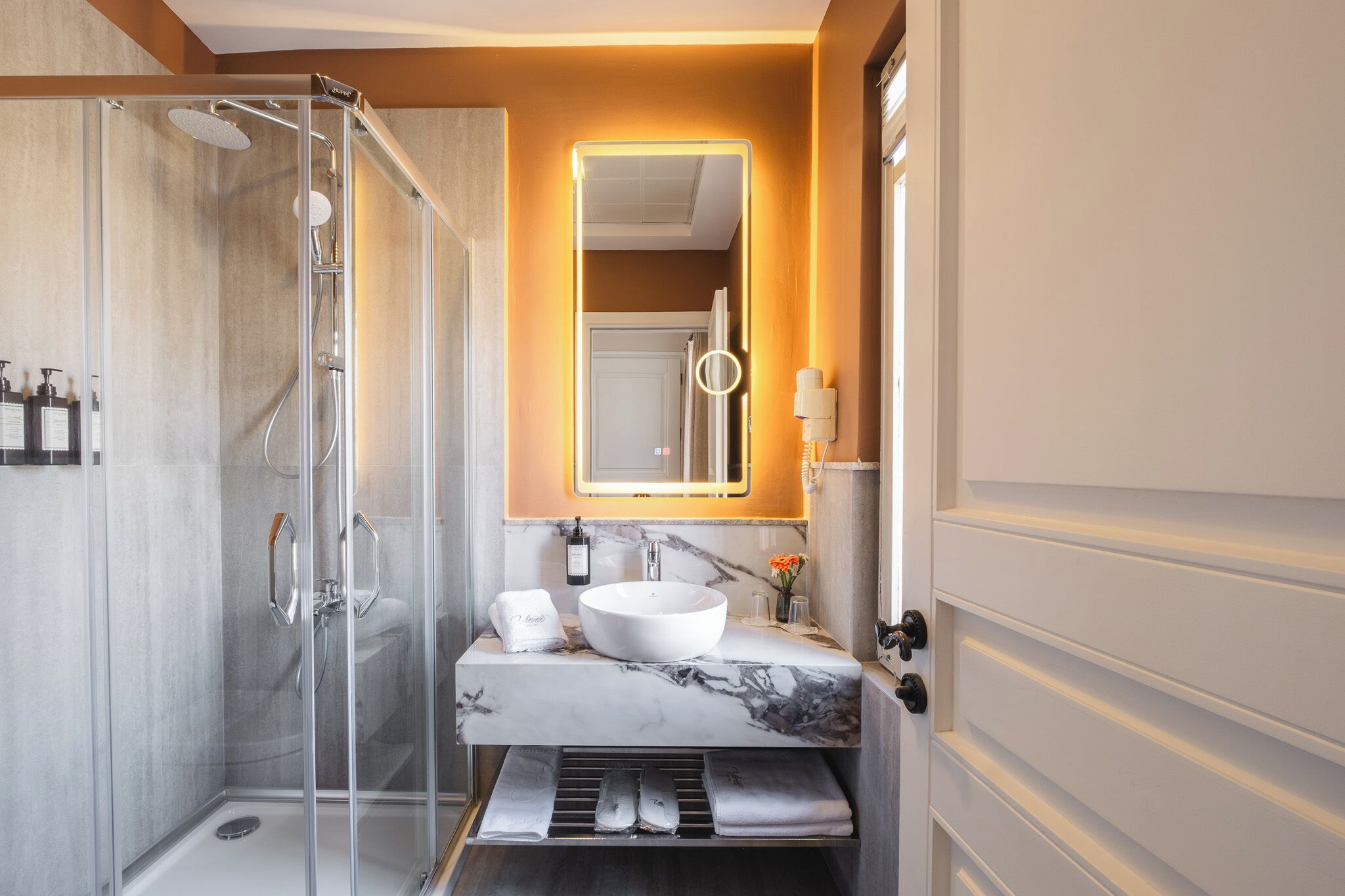 A modern bathroom with a glass-enclosed shower, a marble sink, and a mirror with integrated lighting.