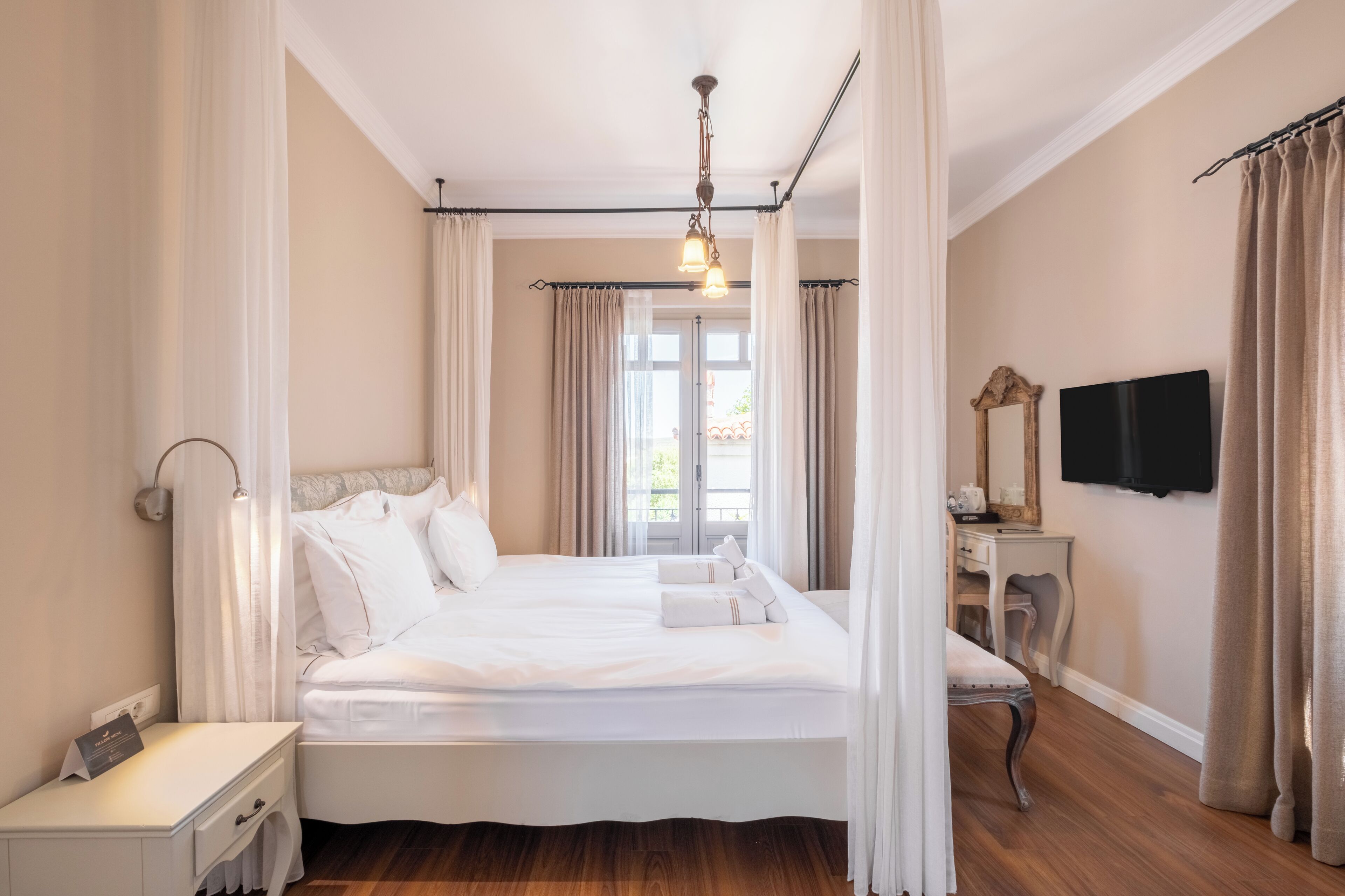 Ein geräumiges Schlafzimmer mit einem großen Bett, einem Nachttisch, einem Schreibtisch, einem Stuhl, einem Fernseher und einem Fenster mit Vorhängen.