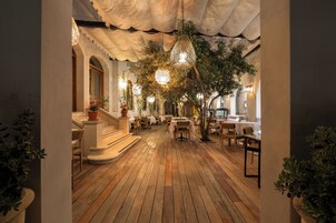 Un comedor interior con piso de madera, mesas, sillas, lámparas colgantes y plantas en macetas.