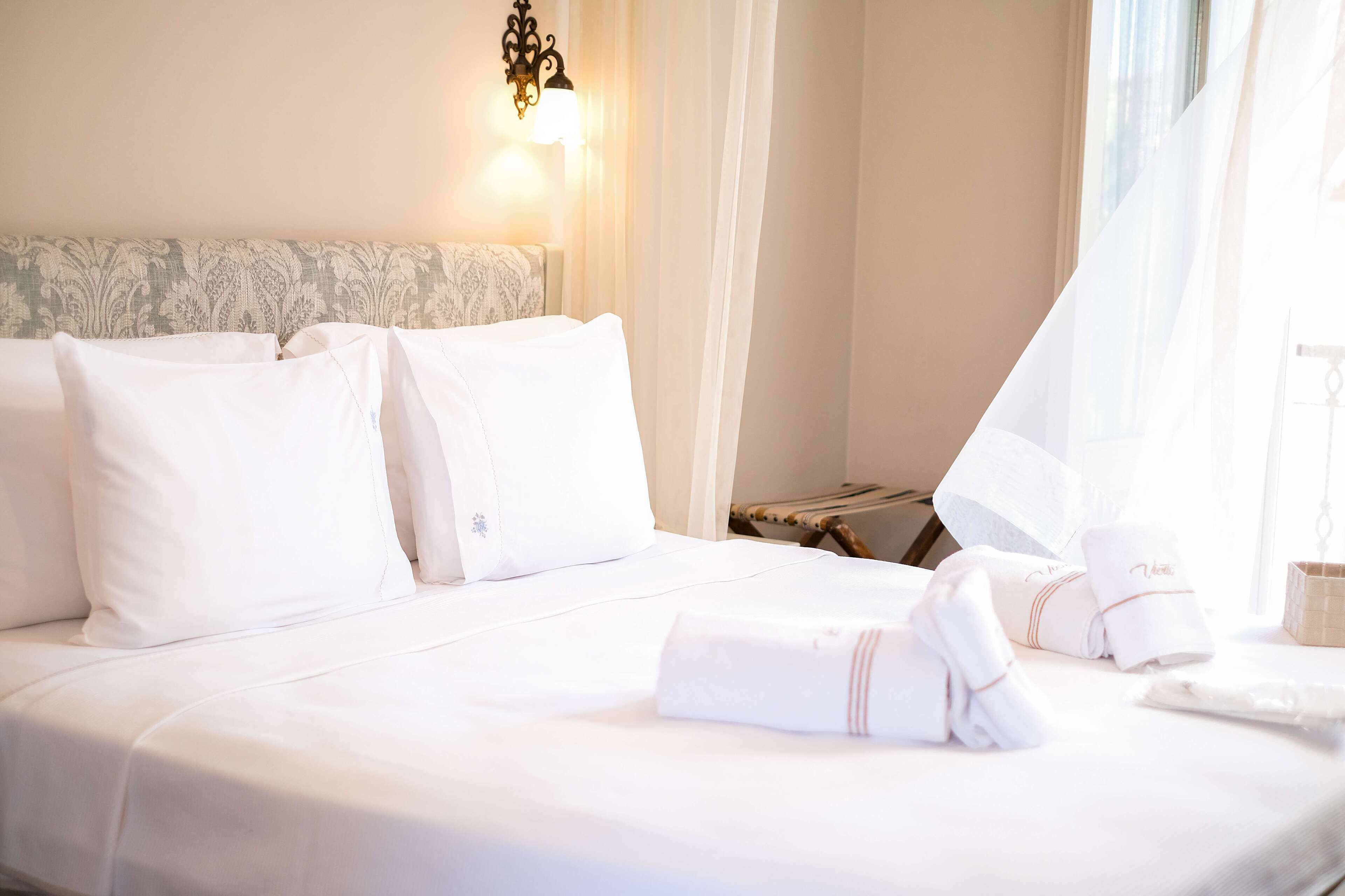 A neatly made bed with white linens, a bedside lamp, and a window with sheer curtains.