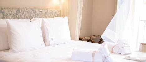 A neatly made bed with white linens, a bedside lamp, and a window with sheer curtains.
