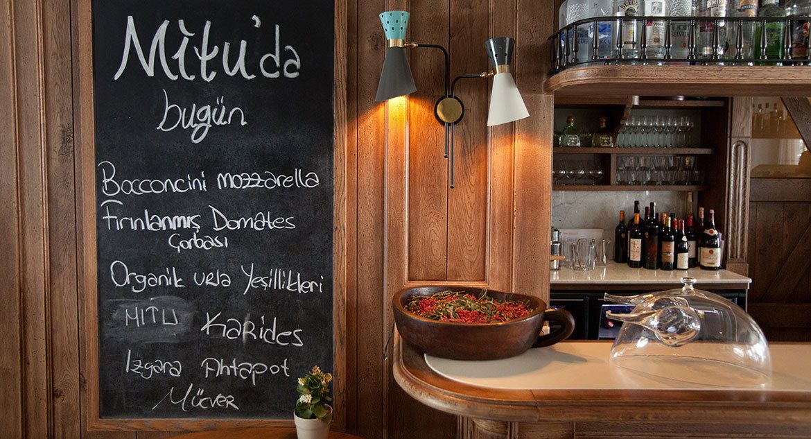 En restauranginteriör med en meny på en svart tavla där rätterna listas, en bardisk i trä med flaskor och en skål med blommor på disken.