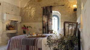 A rustic bedroom with a stone wall, a wooden bed, a window with curtains, and a bedside table with a vase of flowers.