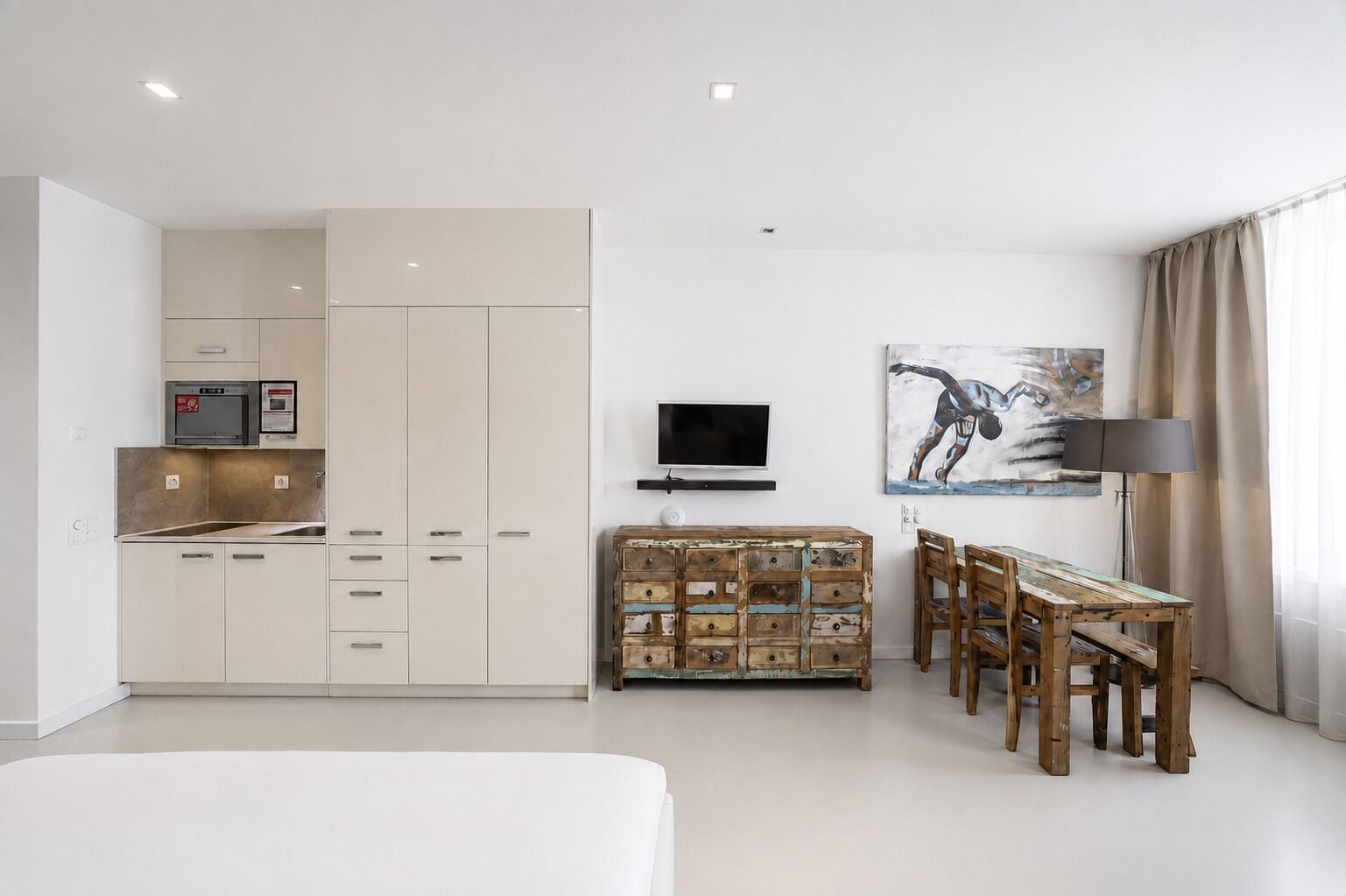 A modern bedroom with a bed, a dining table with chairs, a TV, and a kitchenette.