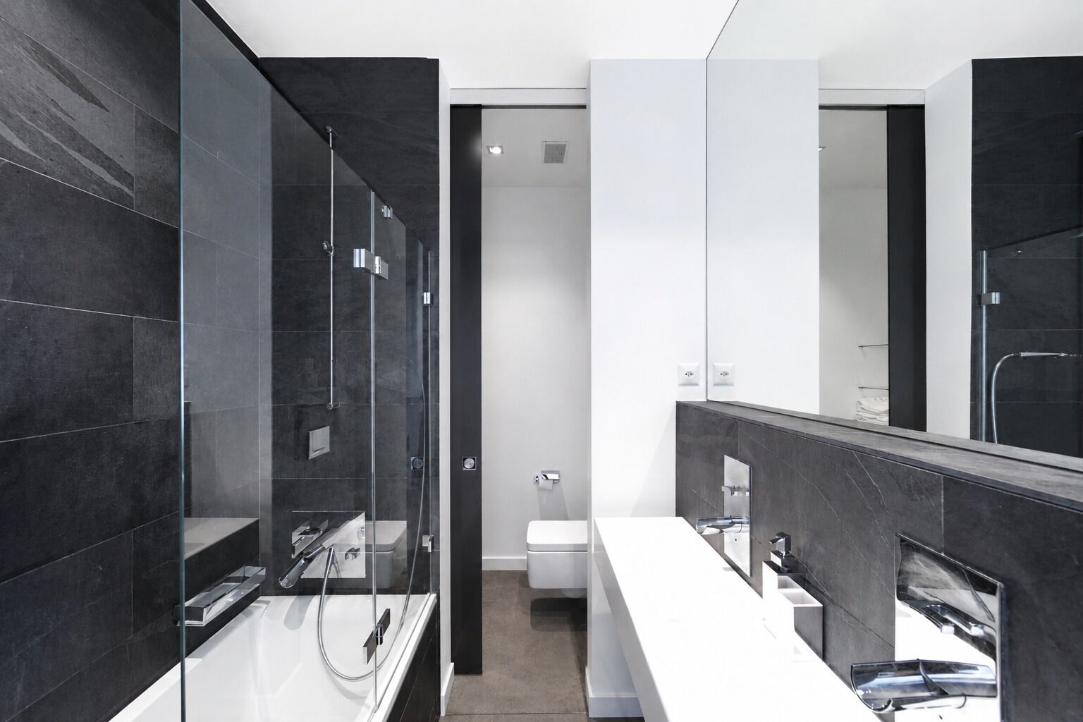 Un bany modern amb parets de rajola fosca, una cabina de dutxa de vidre i un moble de bany blanc amb mirall.