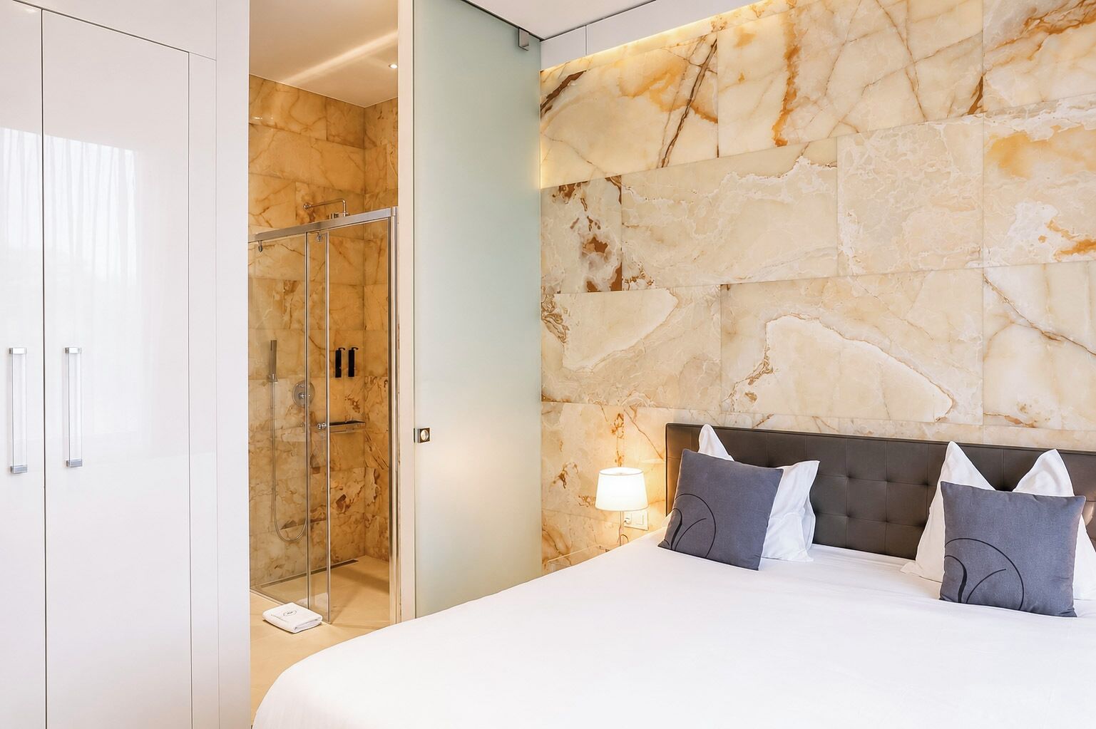 A modern hotel room with a marble wall, a glass-enclosed shower, a bed with a headboard, and a nightstand with a lamp.