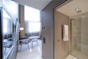Una habitación de hotel moderna con ducha de vidrio, zona de comedor con mesa y sillas, y un amplio ventanal con cortinas.