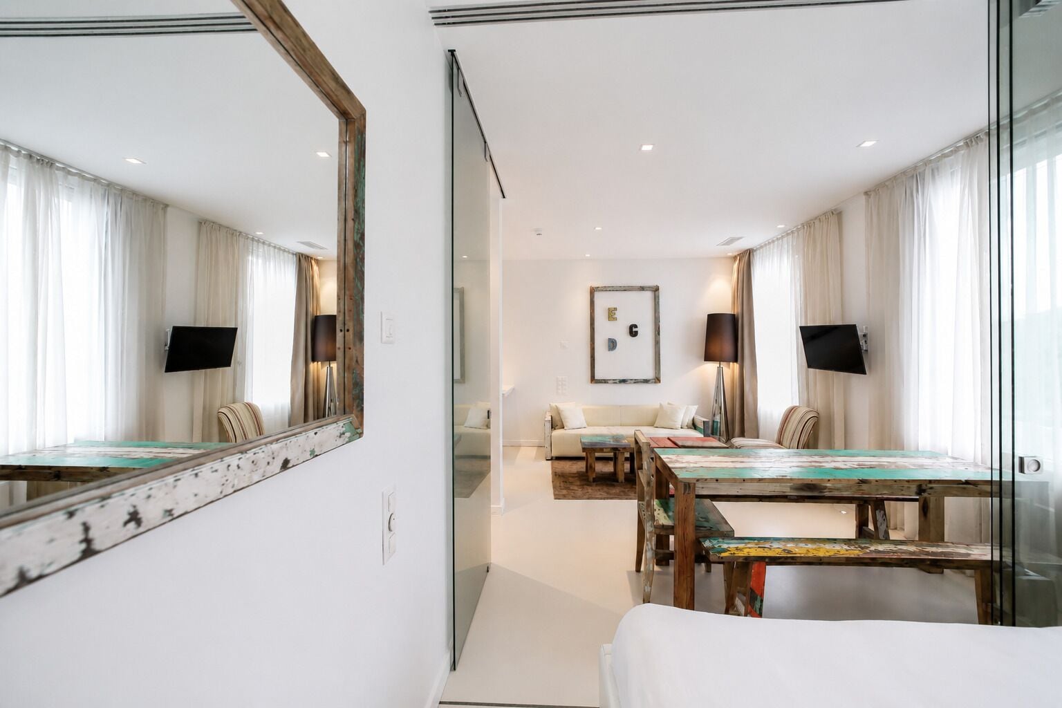 A hotel room with a large mirror, a dining table, a sofa, and a TV mounted on the wall.