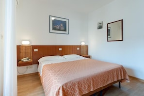 Una cama individual con colcha estampada, cabecera de madera, lámparas de noche, mesita con teléfono y un cuadro en la pared.