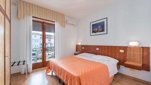 A hotel room with a bed, a wooden headboard, a nightstand, a wall-mounted shelf, and a window with a view of buildings. - Hotel Helvetia (Grado)