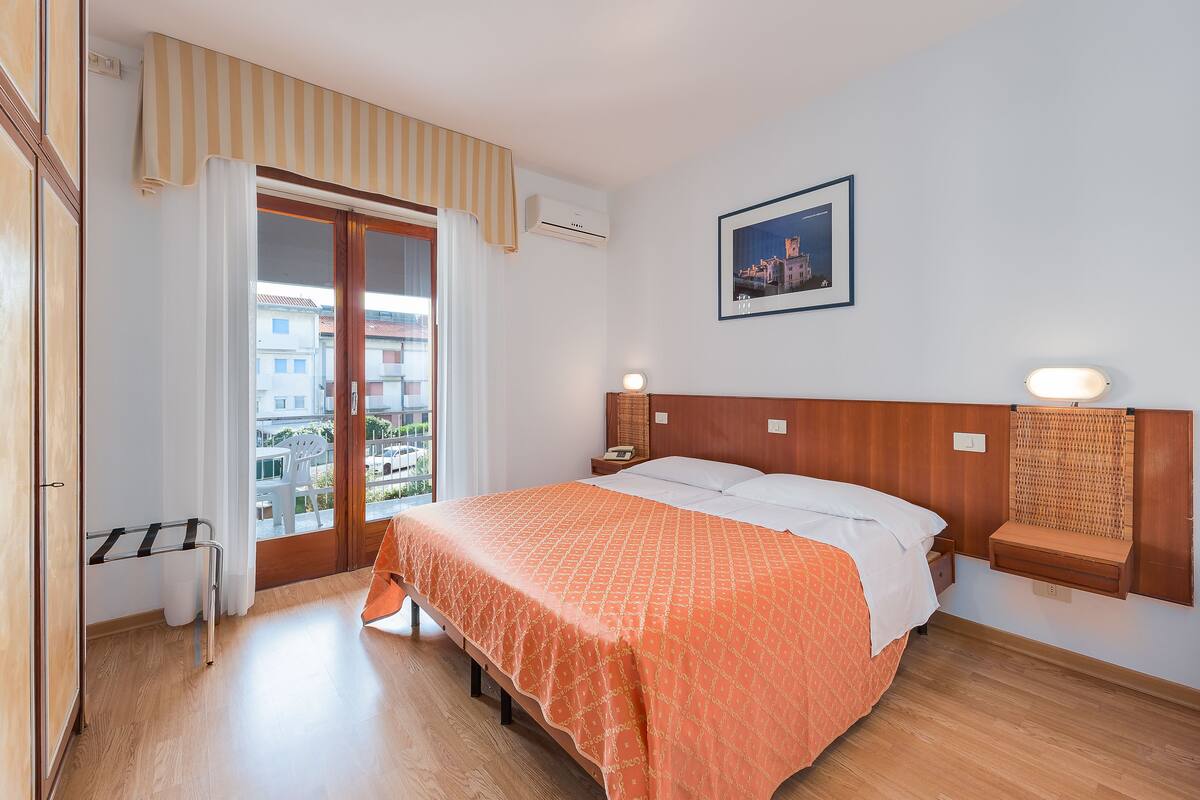 A hotel room with a bed, a wooden headboard, a nightstand, a wall-mounted shelf, and a window with a view of buildings.