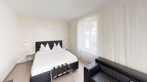 Una habitación de hotel moderna con una cama grande, una mesita de noche, una silla y una ventana con cortinas traslúcidas.