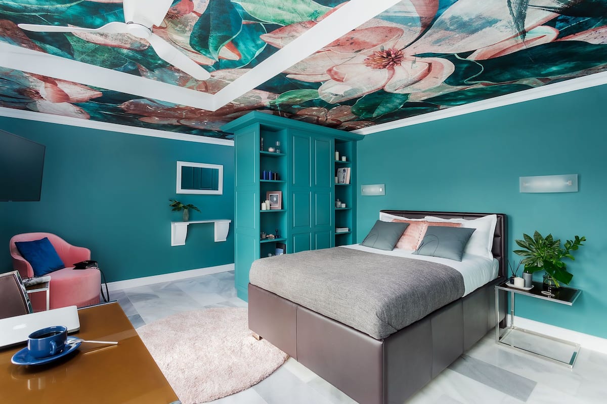 A teal-themed bedroom with a bed, a pink chair, a bookshelf, and a ceiling mural.