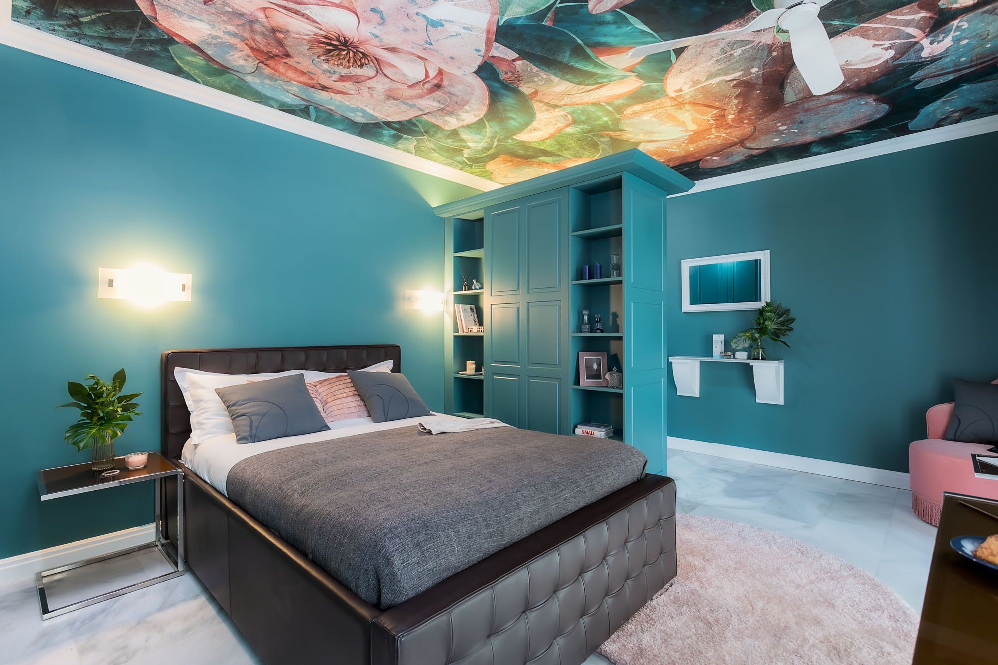 Un dormitorio con paredes color turquesa, una cama con cabecera oscura, un mural en el techo y una estantería empotrada.