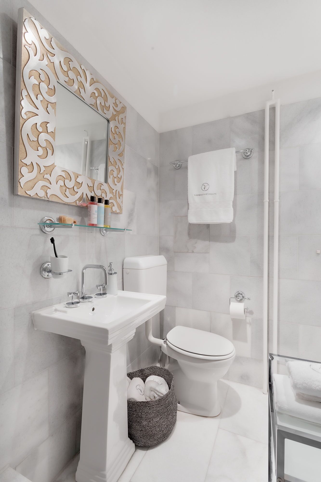 A modern bathroom with a white sink, toilet, and shower. There is a decorative mirror with a gold frame, and toiletries are visible on the sink.