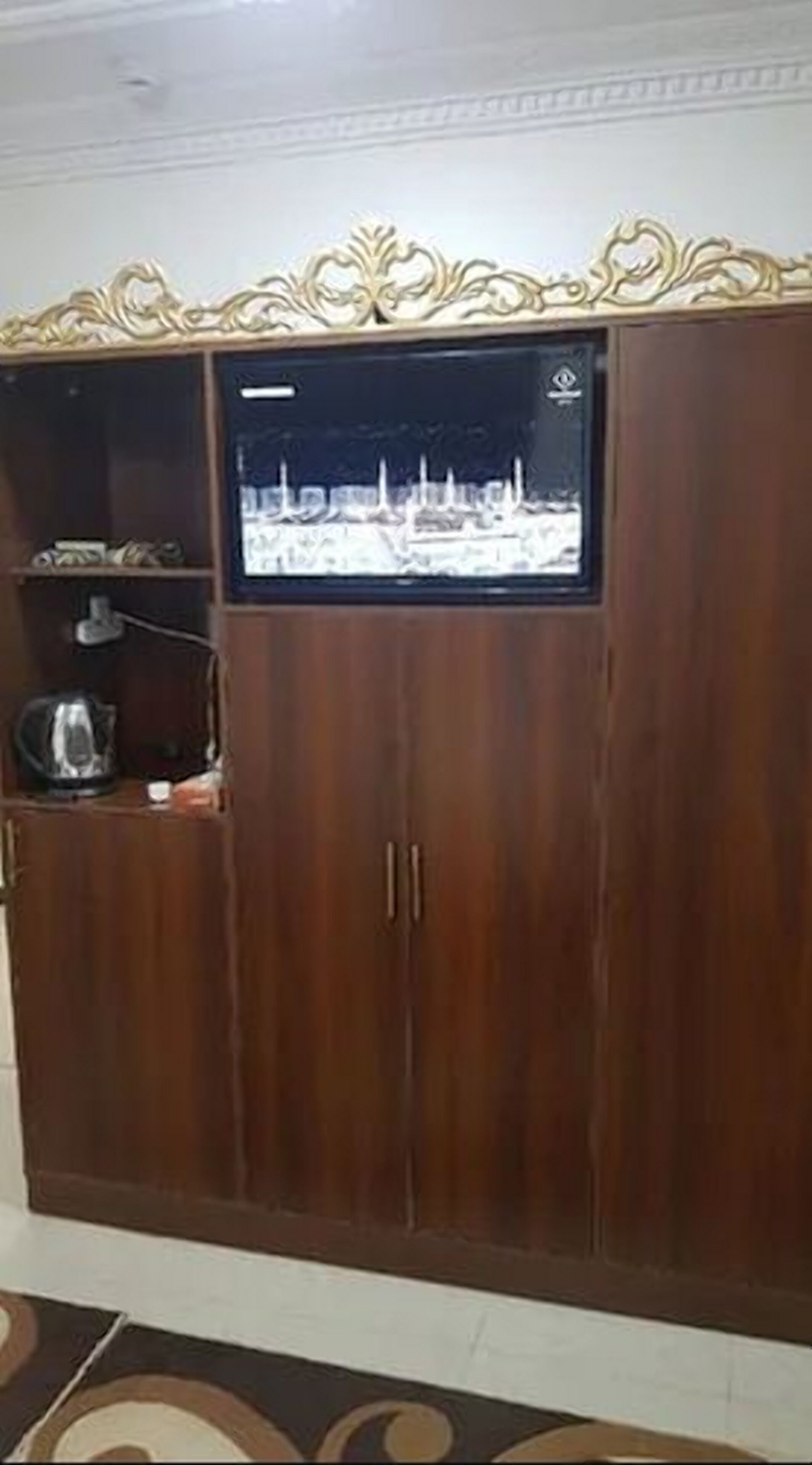 Suuri, koristeellisella päällä varustettu puinen vaatekaappi, jonka sisään on upotettu televisioruutu, jolla näkyy kaupunkimaisema.