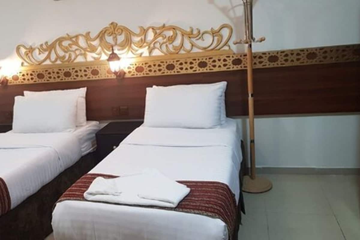 A hotel room with two single beds, each with white linens and a brown headboard. There is a floor lamp and a wall-mounted light fixture.