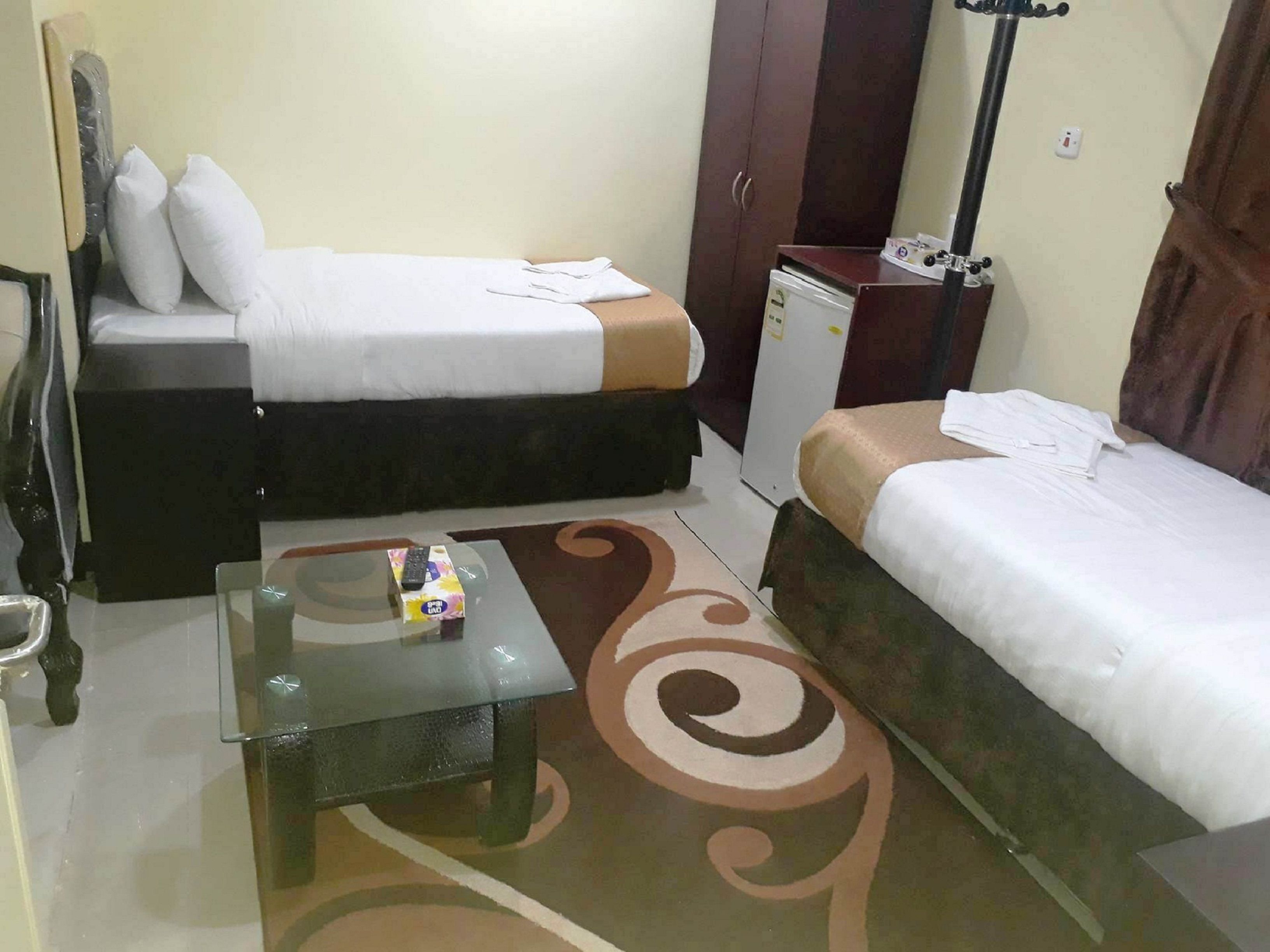 A small hotel room with two beds, a small table, a wardrobe, and a refrigerator.
