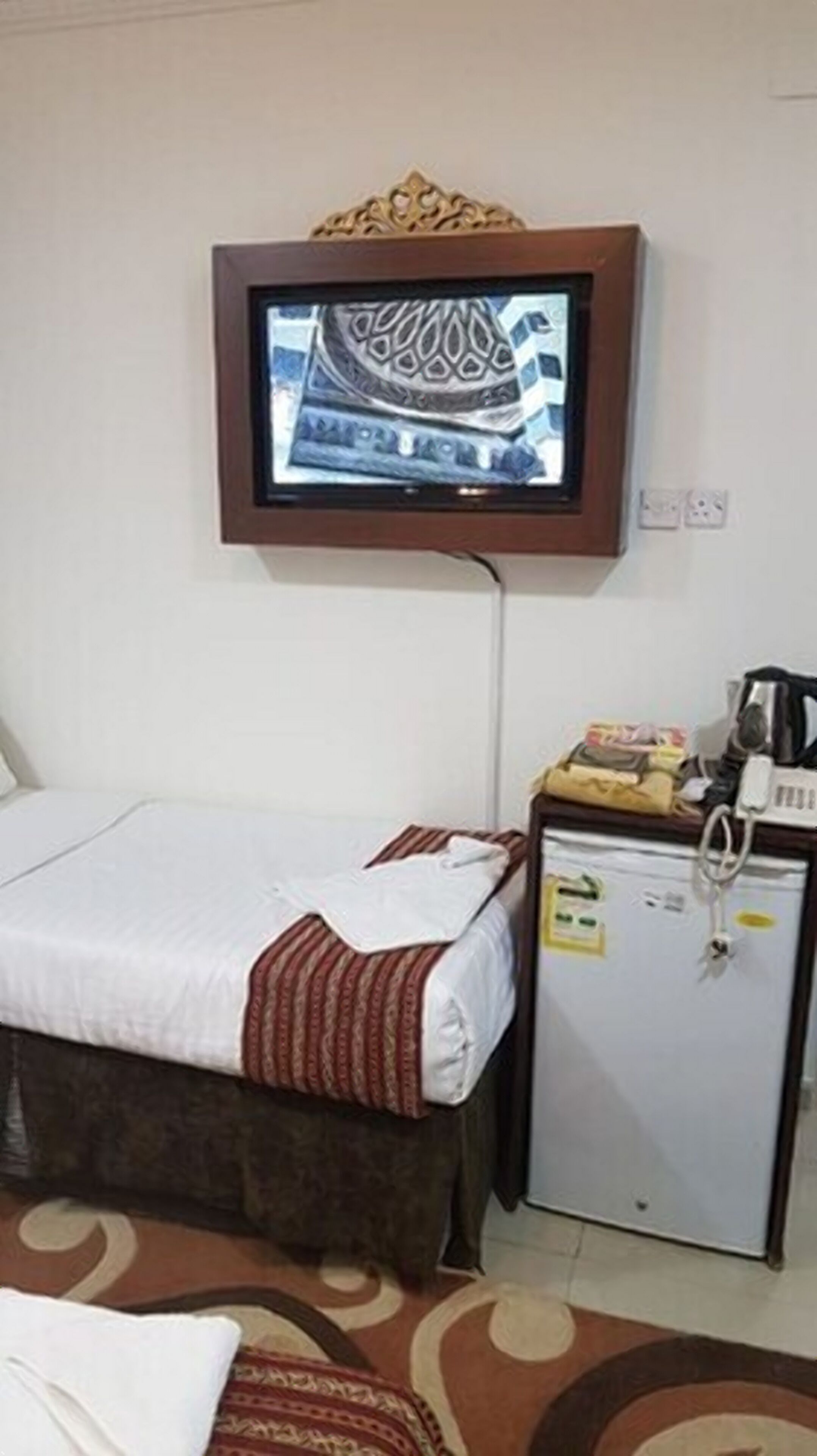 A hotel room with a bed, a television mounted on the wall, a small refrigerator, and a bedside table with a kettle.
