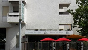 A modern building with multiple balconies and a ground-level restaurant with red umbrellas.