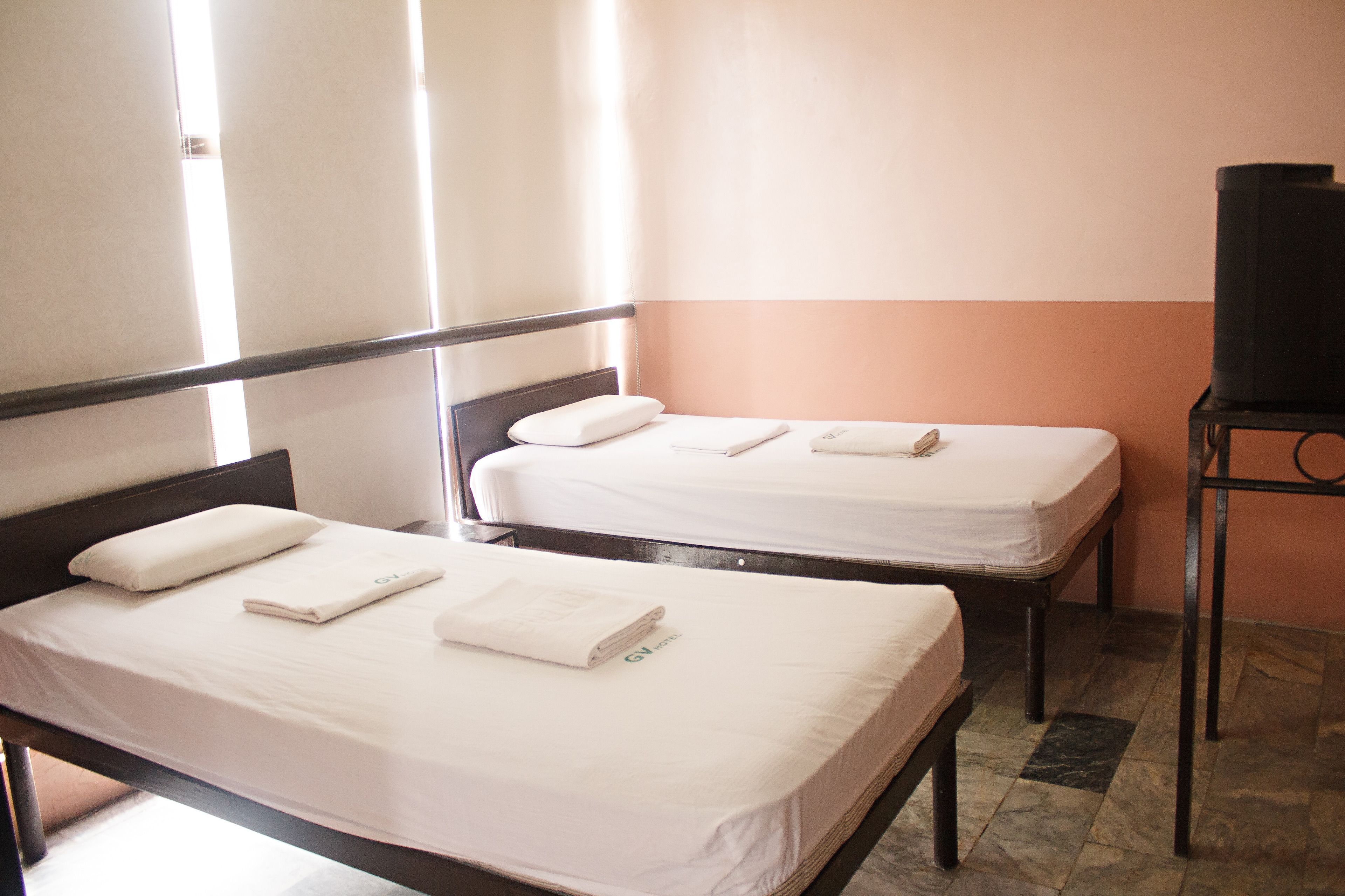 Two single beds with white linens and pillows, a television on a stand, and a tiled floor.